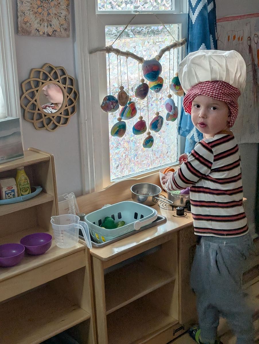A child wearing a pink knitted hat plays at a wooden play kitchen.